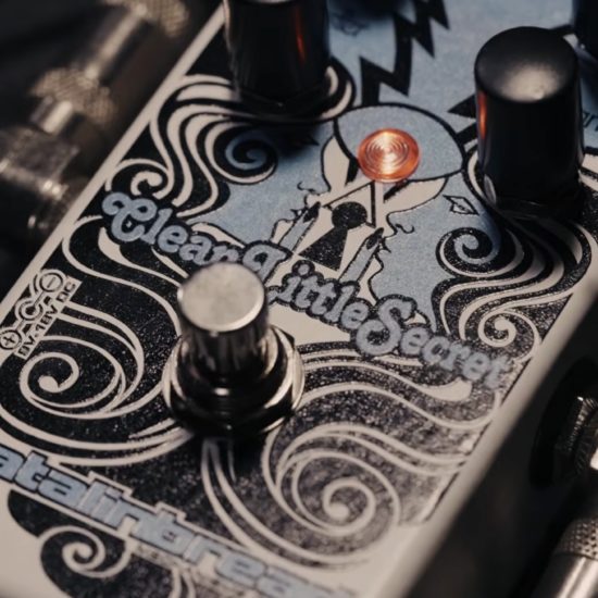 Close-up of a Catalinbread Clean Little Secret guitar effects pedal with decorative artwork, glowing LED indicator, footswitch, and control knobs, connected with instrument cables.